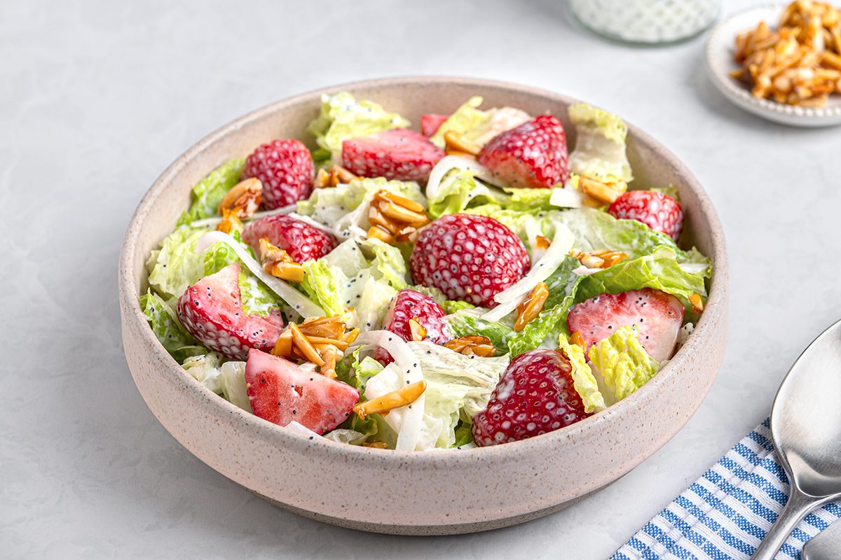 A bowl of salad featuring romaine lettuce, fresh strawberries, sliced almonds, and a sprinkle of shredded cheese. The bowl is placed on a light gray surface, with a blue and white striped napkin and spoon nearby.