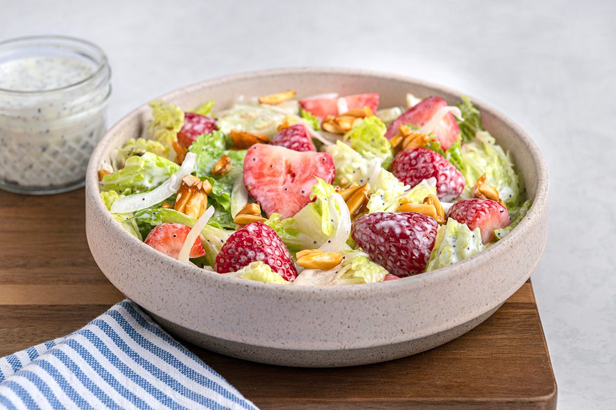 A salad bowl with strawberries, lettuce, sliced almonds, and creamy dressing. A small glass jar of extra dressing is beside it on a wooden surface, with a blue and white striped cloth nearby.