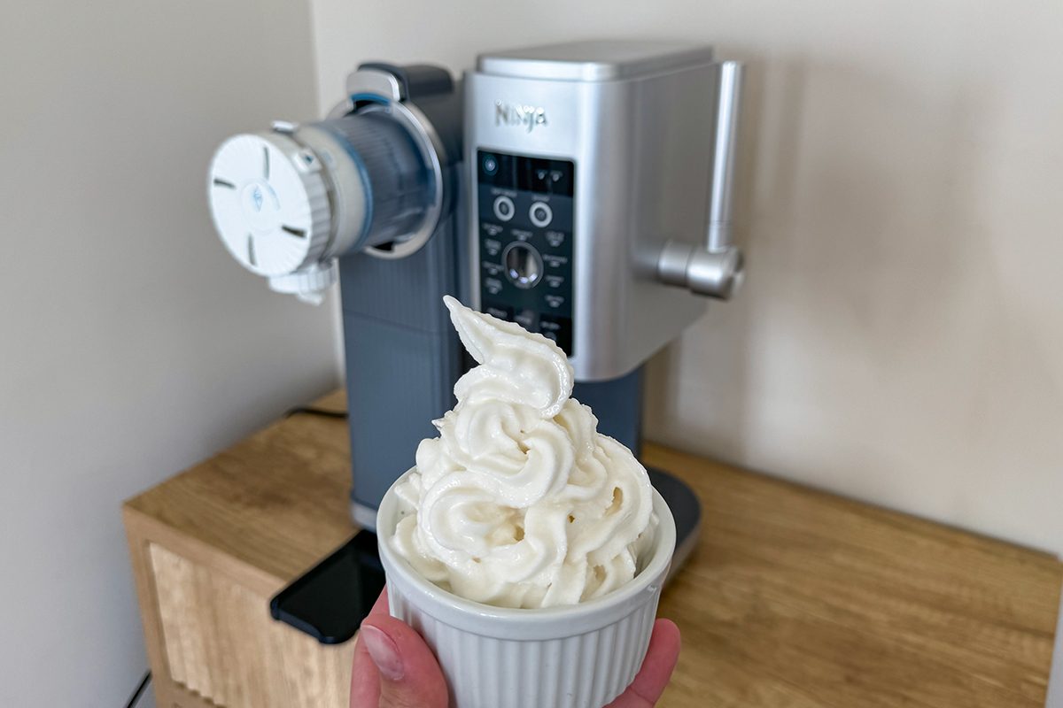 A person holds a white ceramic bowl filled with soft serve ice cream in front of a silver Ninja ice cream maker on a wooden countertop. The ice cream is swirled high above the rim of the bowl.