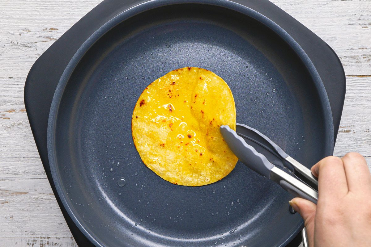 overhead shot of a person using tongs to flip a yellow corn tortilla in a black skillet, the skillet is placed on a white wooden surface