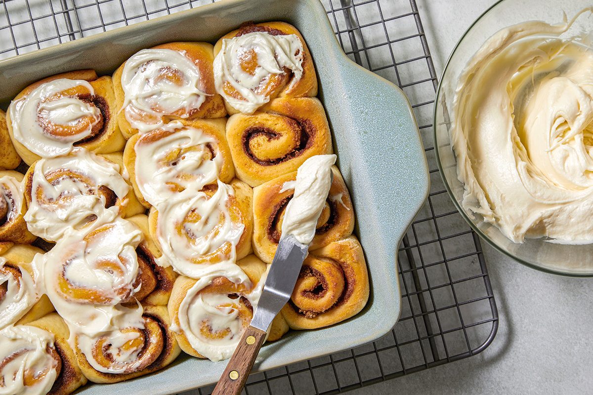 Cinnamon rolls with creamy frosting in a rectangular baking dish on a cooling rack. One roll is being frosted with a spatula. A bowl of additional frosting is visible on the side.