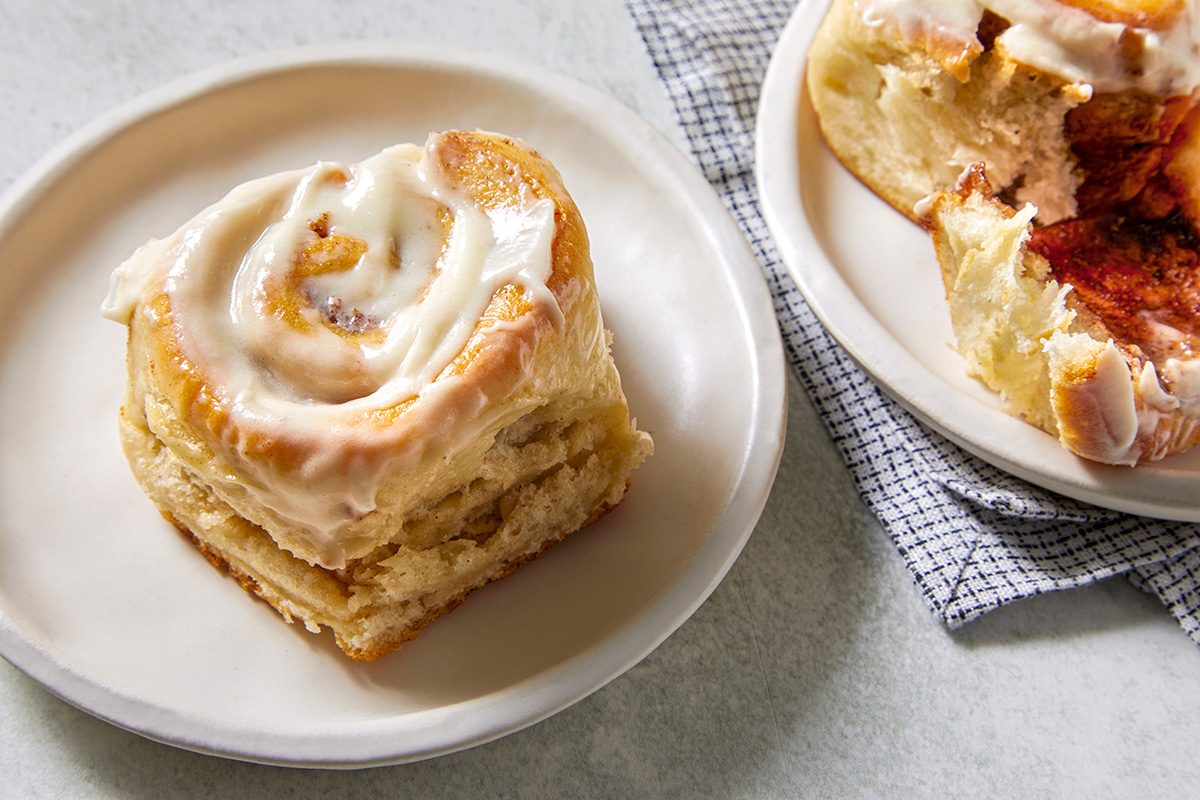 A close-up of a frosted cinnamon roll on a white plate, sitting on a light-colored table. Another cinnamon roll is partially visible on a second plate nearby, with a checkered cloth underneath. The rolls are topped with a creamy glaze.