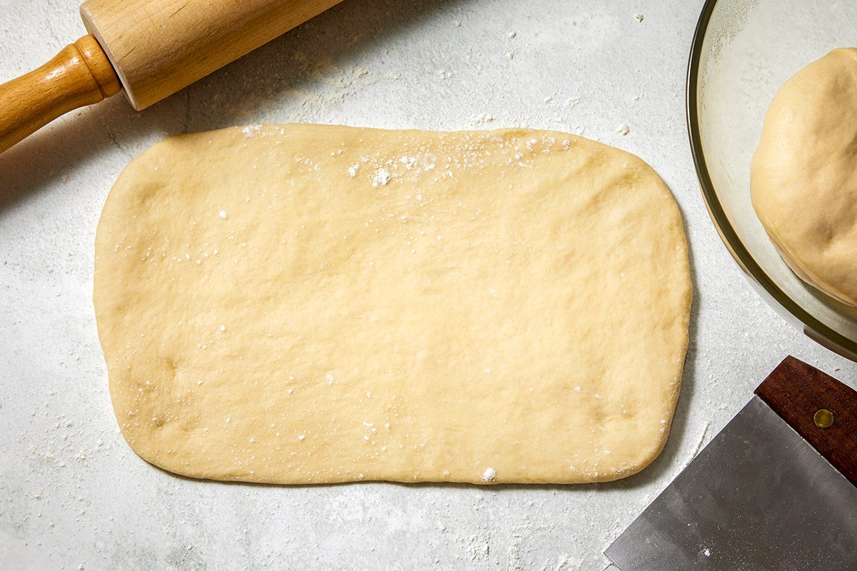 Flattened dough on a lightly floured surface, with a rolling pin nearby. A ball of dough and a dough scraper are visible in the background.