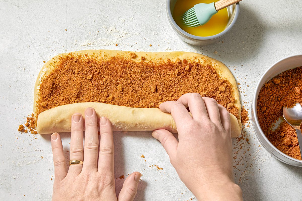 Hands rolling dough with a cinnamon and sugar mixture on a light surface. A bowl of melted butter with a brush and a bowl with remaining cinnamon sugar are nearby.