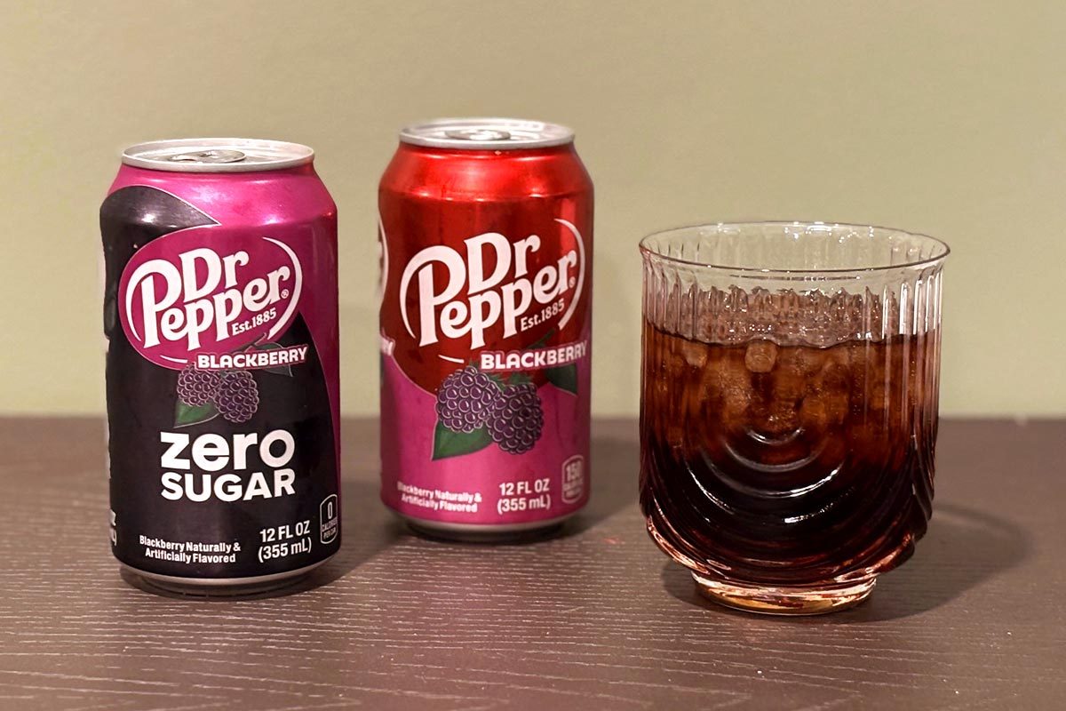 Can of Dr Pepper Blackberry Zero Sugar and a can of regular Blackberry Dr Pepper, next to a glass of the soda with ice, on a wooden table with a beige background