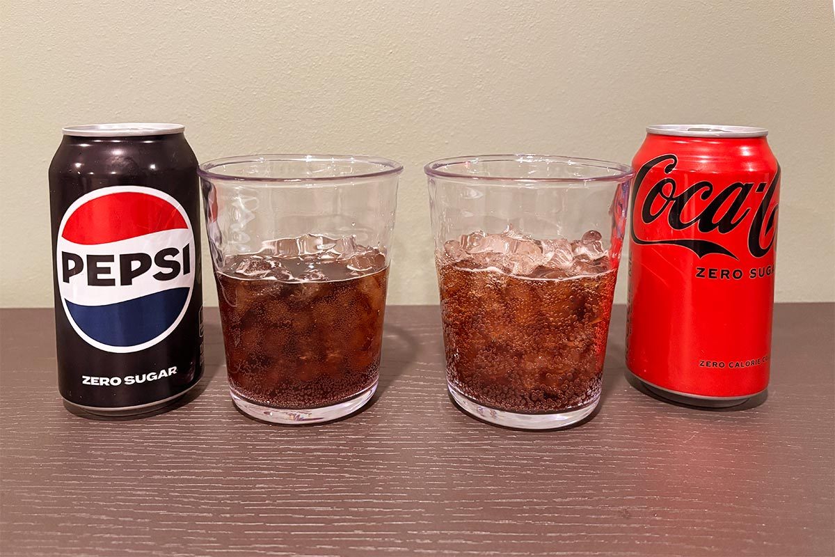 Black can of Pepsi Zero sugar with a glass of pepsi zero over ice on the left, with a Red can of Coca Cola Zero Sugar next to a glass of coca cola zero on ice on the right, both on a wooden table and beige background