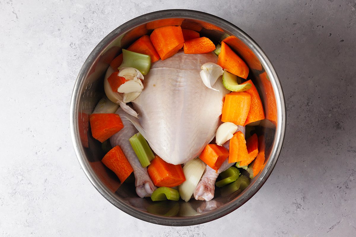 A raw chicken lies in a metal bowl surrounded by chopped carrots, celery, onions, and garlic cloves, ready for cooking. The ingredients are placed on a light gray surface.
