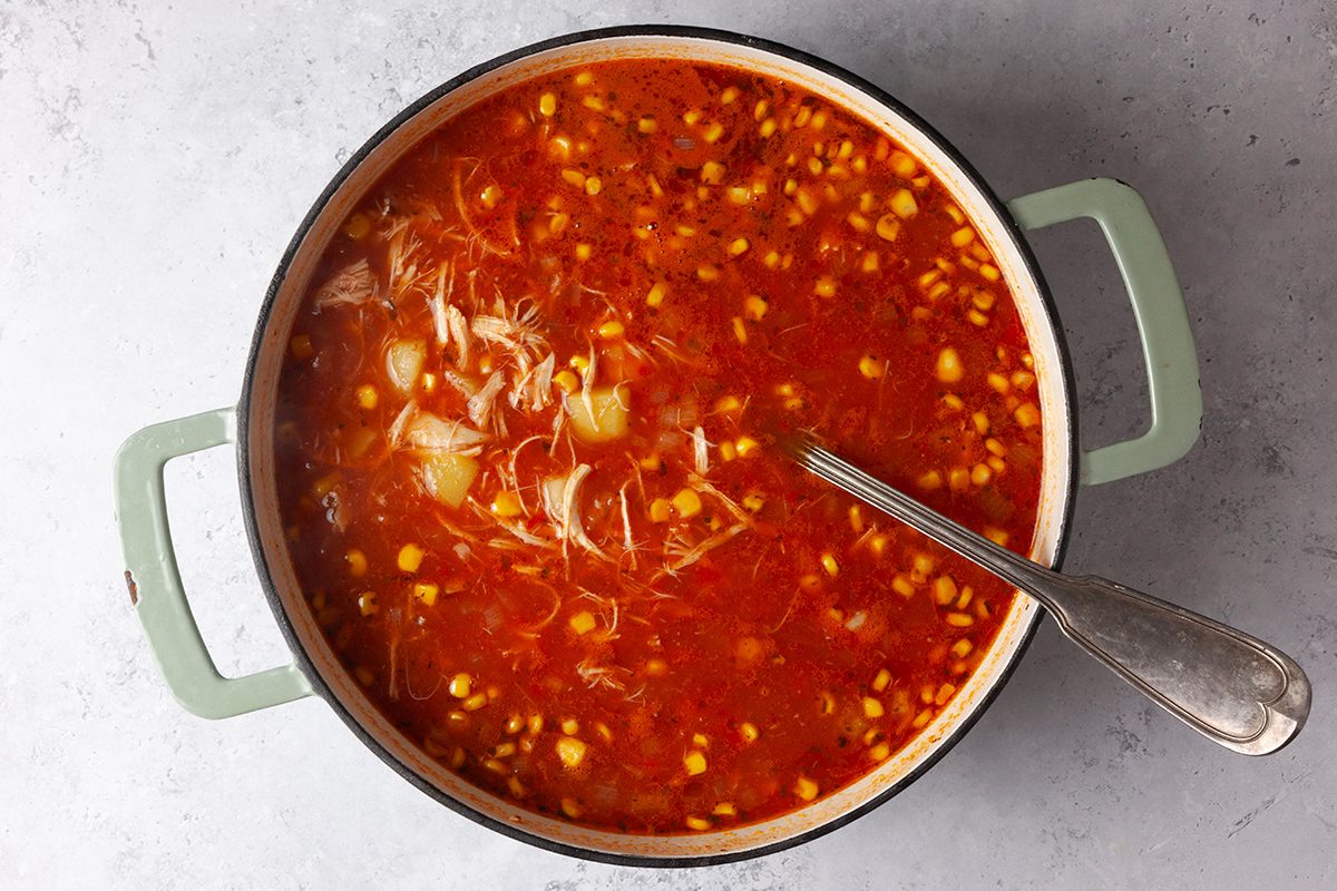A pot of red soup filled with shredded chicken, corn, and onions. A spoon rests inside the pot, and the handles are light green. The background is a speckled, light gray surface.