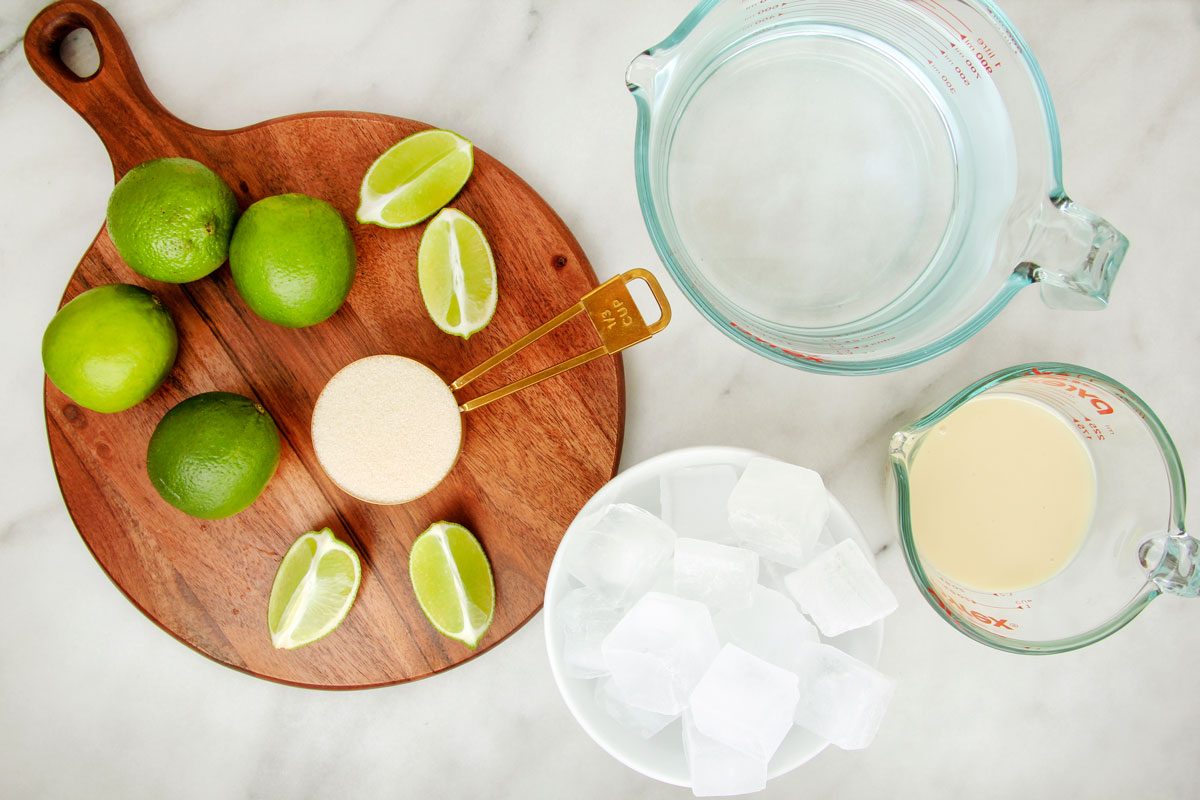 A top view of ingredients for Brazilian Lemonade on a marble surface: a wooden board with whole and sliced limes, a glass of a creamy drink, a measuring tool, a bowl of ice cubes, and two measuring cups with liquid.