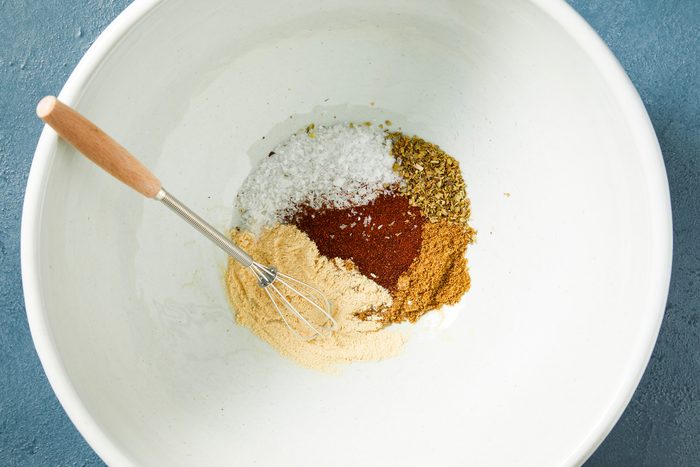 Overheda shot of a large bowl combine salt; cumin, garlic powder, chili powder and oregano; whisk tool; dark blue surface;