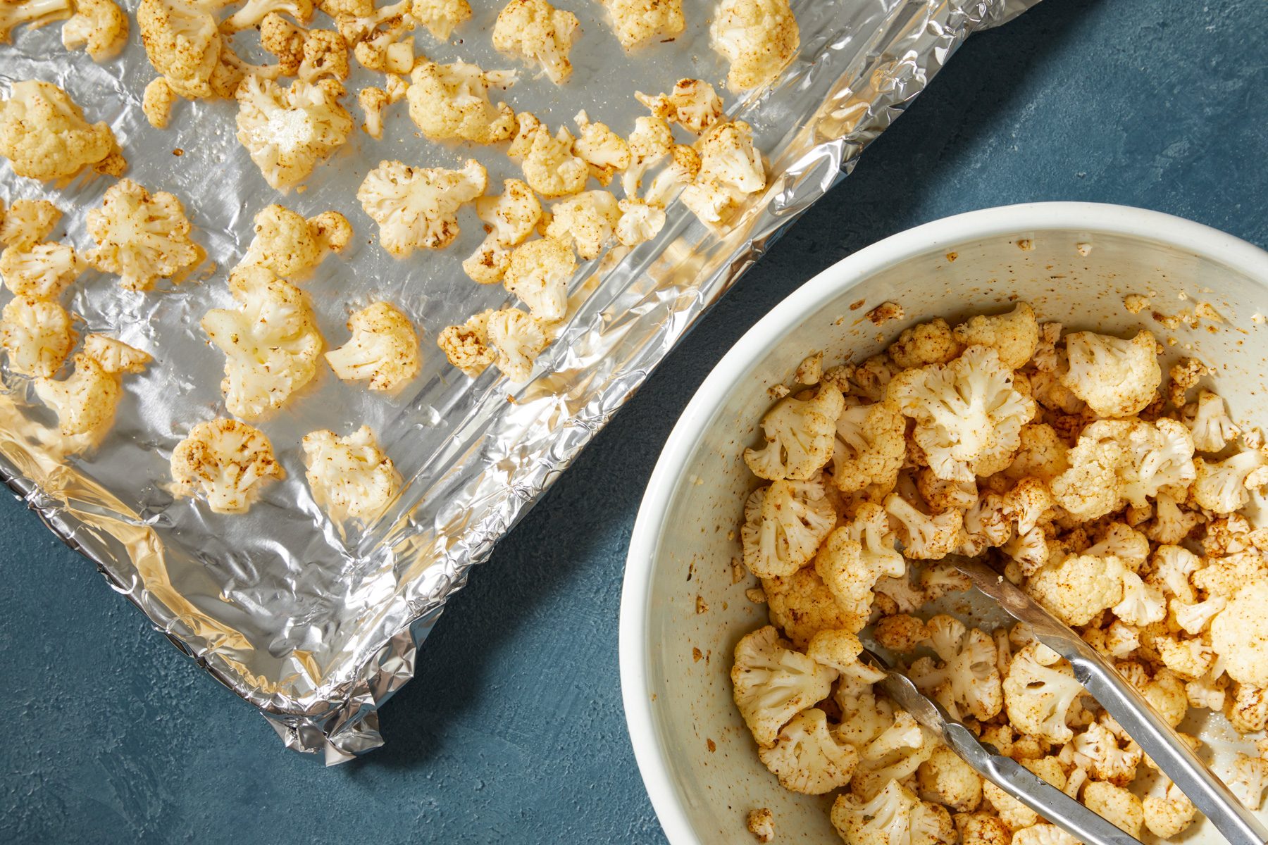 Overhead shot of add cauliflower and oil into mixture; toss to coat; Transfer to a foil-lined baking sheet; tongs; dark blue surface;