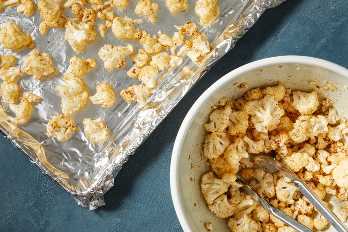 Overhead shot of add cauliflower and oil into mixture; toss to coat; Transfer to a foil-lined baking sheet; tongs; dark blue surface;