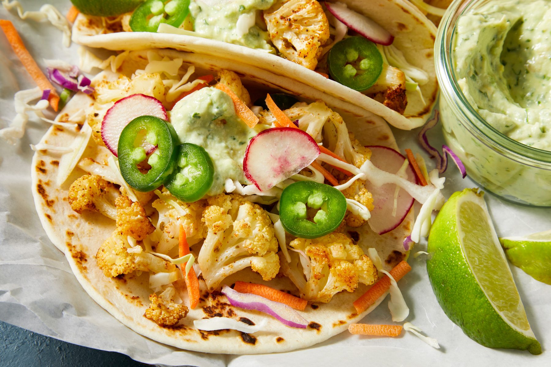 Close-up shot of Cauliflower Tacos; Serve in tortillas with coleslaw mix avocado crema and toppings; lime wedges; dark blue surface;