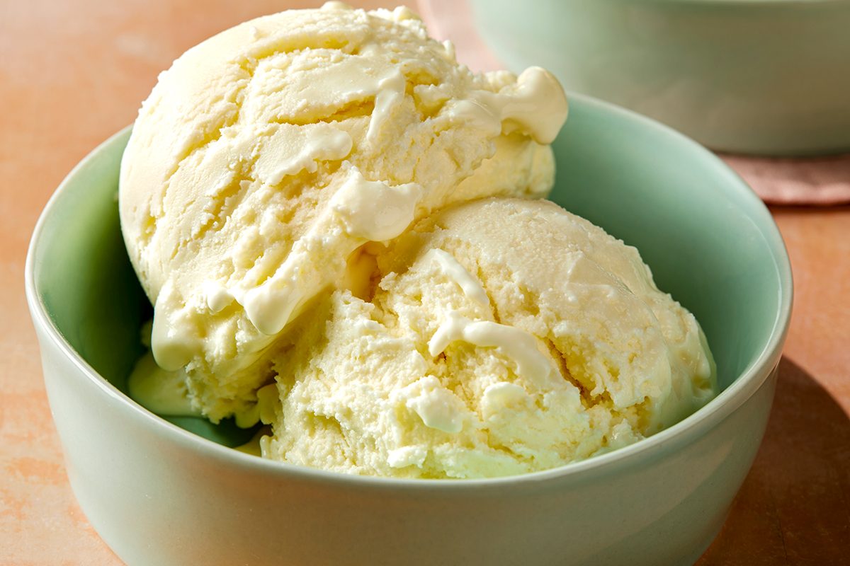 A bowl containing two generous scoops of creamy vanilla ice cream, displayed in a light green bowl on a warm-toned surface.