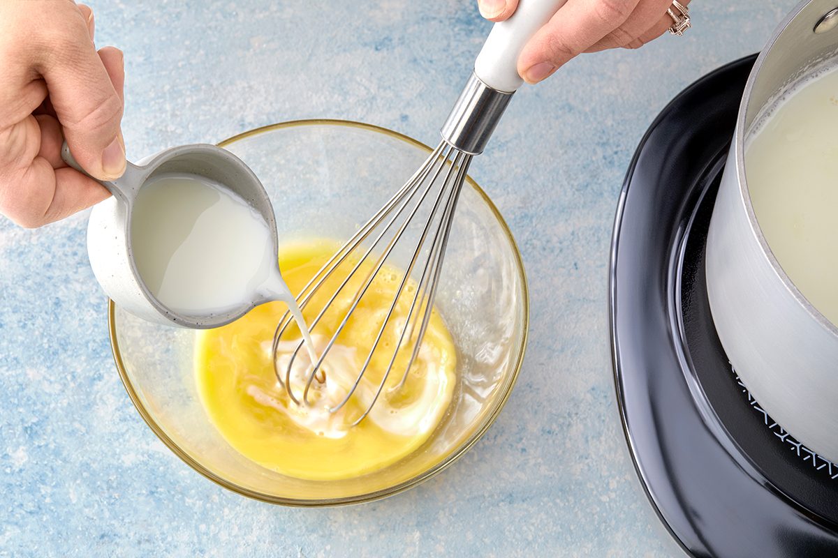 A person pours milk from a small jug into a glass bowl containing yellow beaten eggs. A whisk is also in the bowl. A saucepan with a light liquid inside is on a stove nearby. The background is a light blue surface.