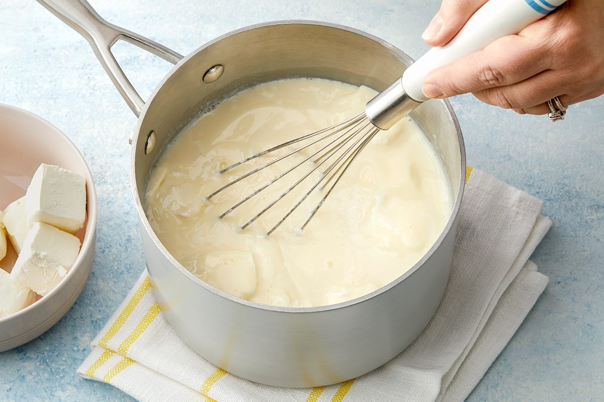 Hand whisking a creamy white sauce in a saucepan on a stovetop, with a bowl of butter cubes nearby. The saucepan is resting on a folded cloth with yellow stripes on a blue surface.