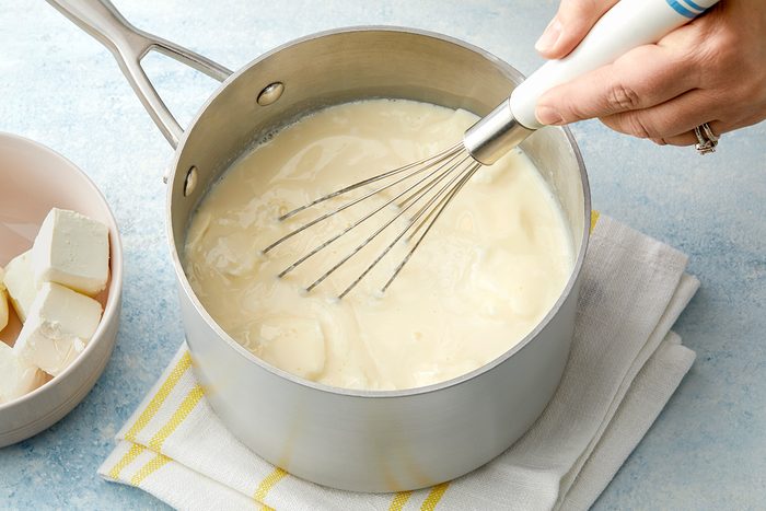 Hand whisking a creamy white sauce in a saucepan on a stovetop, with a bowl of butter cubes nearby. The saucepan is resting on a folded cloth with yellow stripes on a blue surface.