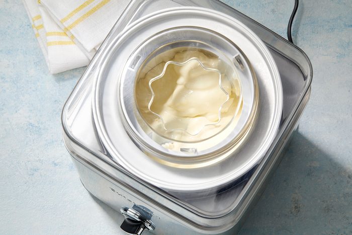 A silver countertop ice cream maker with a transparent lid is shown from above. Inside, vanilla ice cream is being churned. A folded white and yellow striped cloth is placed next to the machine.