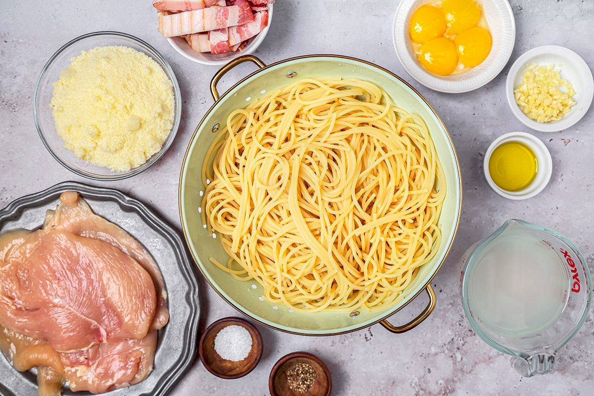 overhead shot of a pasta dish; at the center, there is a colander filled with cooked spaghetti; surrounding the pasta are various components
