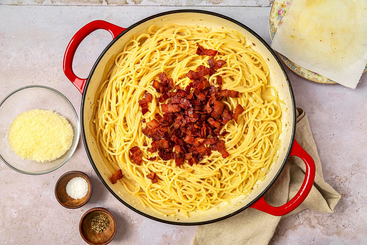 overhead shot of pasta placed in a large, shallow red pot and topped with crispy bits of bacon