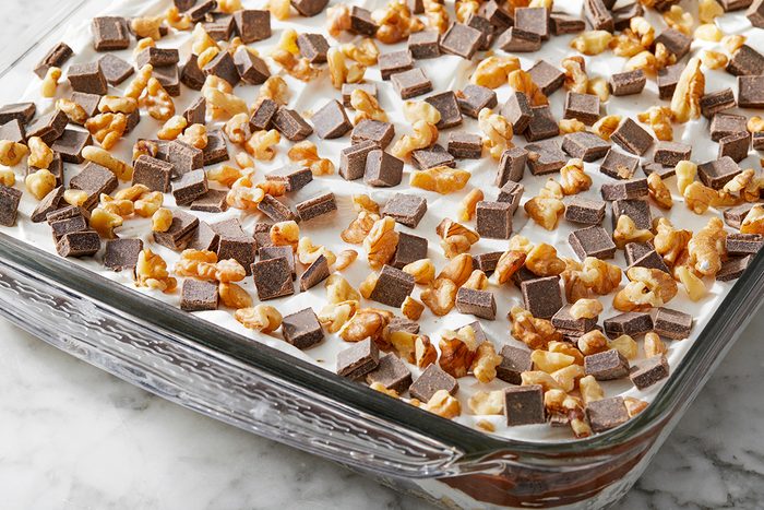 A glass dish filled with a layered dessert topped with chopped walnuts and chocolate pieces. The dessert has visible layers of cream and chocolate, and is placed on a marble surface.