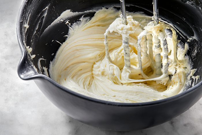 A black mixing bowl filled with creamy, light-colored batter being whipped by an electric mixer. The whisks are coated with the smooth mixture. The bowl is placed on a gray marble surface.
