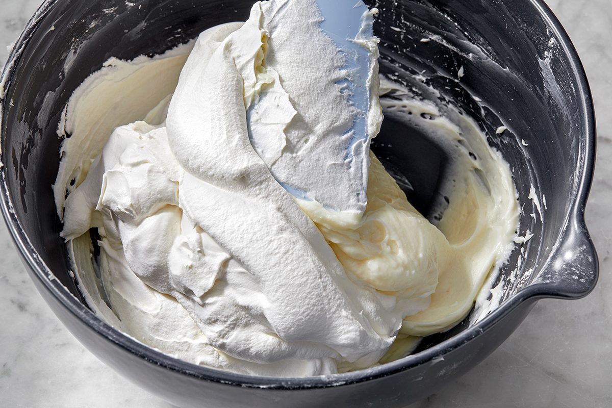 A mixing bowl filled with whipped cream. A metal spatula is partially covered in the cream, lying across the top of the mixture. 