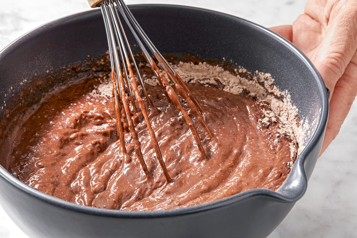 A hand holds a blue mixing bowl containing chocolate batter, with a whisk being used to stir it. The batter is partially mixing with dry ingredients along the edges of the bowl. 