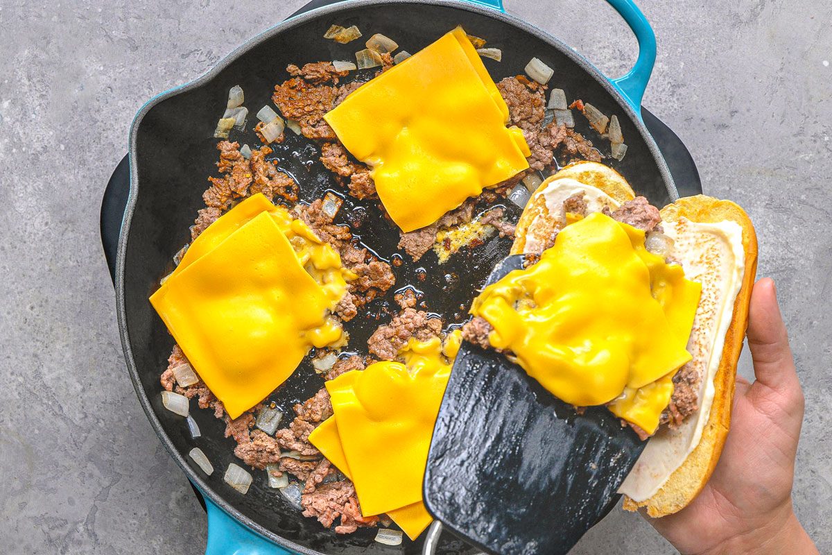 overhead shot of slices of bright yellow cheese being placed on top of cooked ground beef and onions in a black pan, a black spatula is being used to transfer a slice of cheese, and a toasted bun is being held next to the pan, the pan is positioned on a textured grey surface