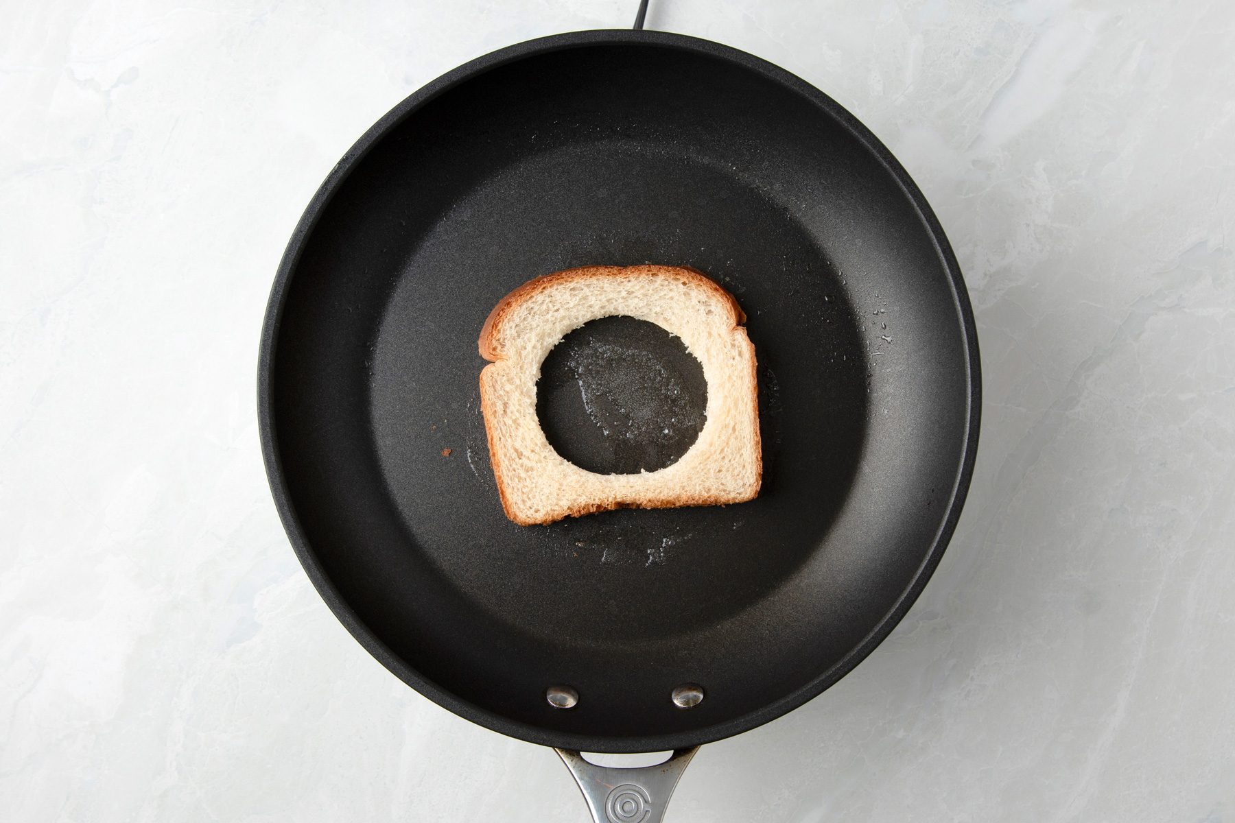 Overhead shot; cut a 3-inch hole in the middle of the bread and discard; In a small skillet melt the butter; place the bread in the skillet; on marble surface;