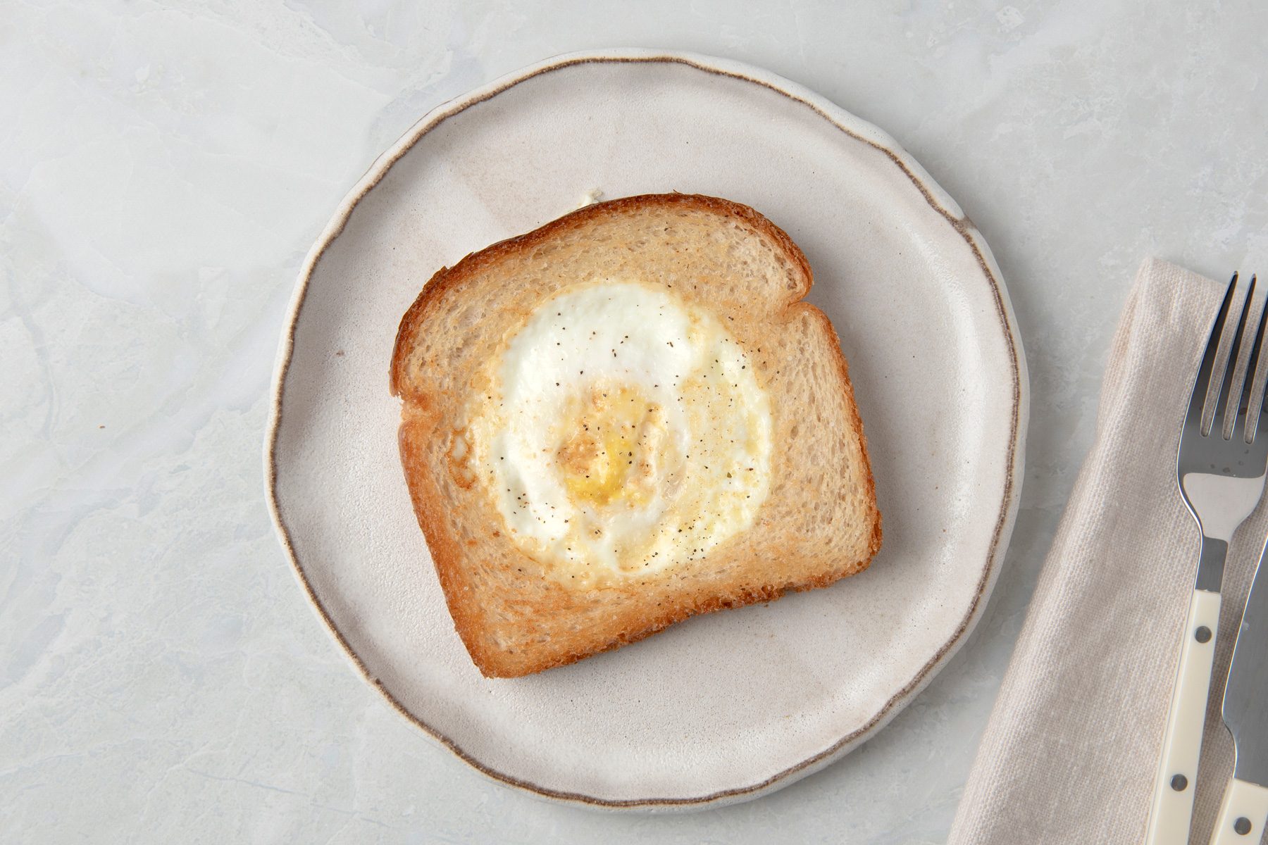 Overhead shot of Toad in the Hole; served ona ceramic plate; season with salt and pepper; fork and knife over a napkin on right side; on a marble surface;