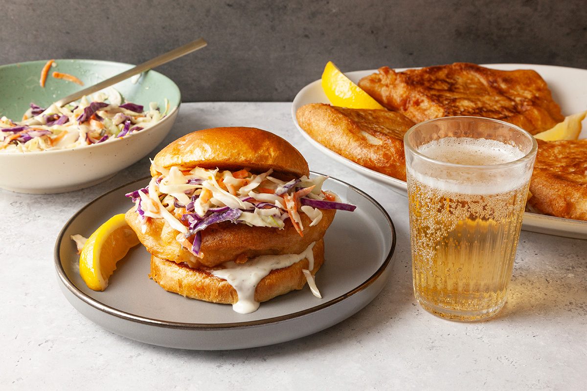 A fish sandwich with coleslaw on a plate, accompanied by a lemon wedge, is set next to a glass of beer. In the background, a bowl of coleslaw and a plate with large fried fish fillets are visible.