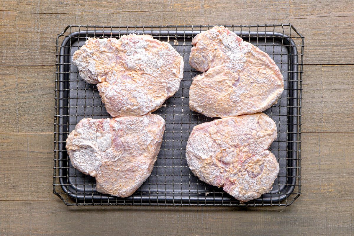 overhead shot of a tray with four pieces of batter coated meat resting on a wire rack, the tray itself has a rectangular shape with a metal design, placed over wooden surface