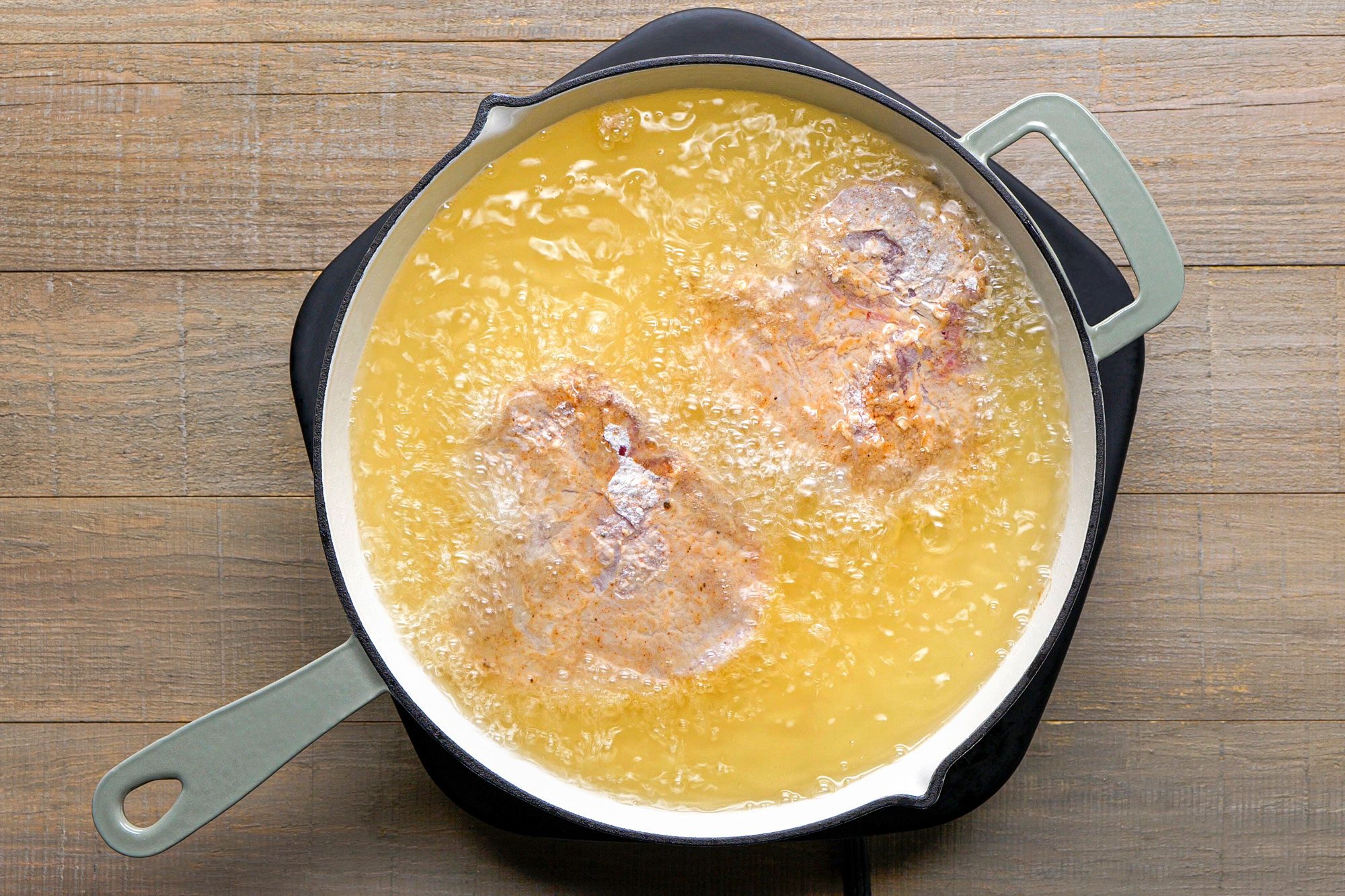 overhead shot of a frying pan filled with hot oil, and two pieces of meat are submerged in the bubbling oil, the background features a wooden surface