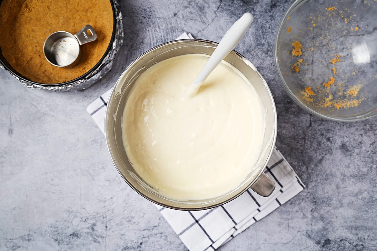 A bowl filled with creamy batter and a spatula sits on a checkered cloth. Nearby, there's a round pan with crushed biscuits and a small measuring cup.