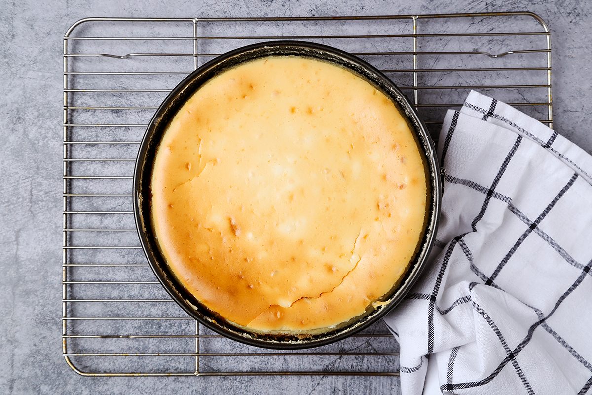 A round cheesecake rests on a cooling rack with a light golden top. A white tea towel with black grid lines is draped near the rack. The background is a textured gray surface.