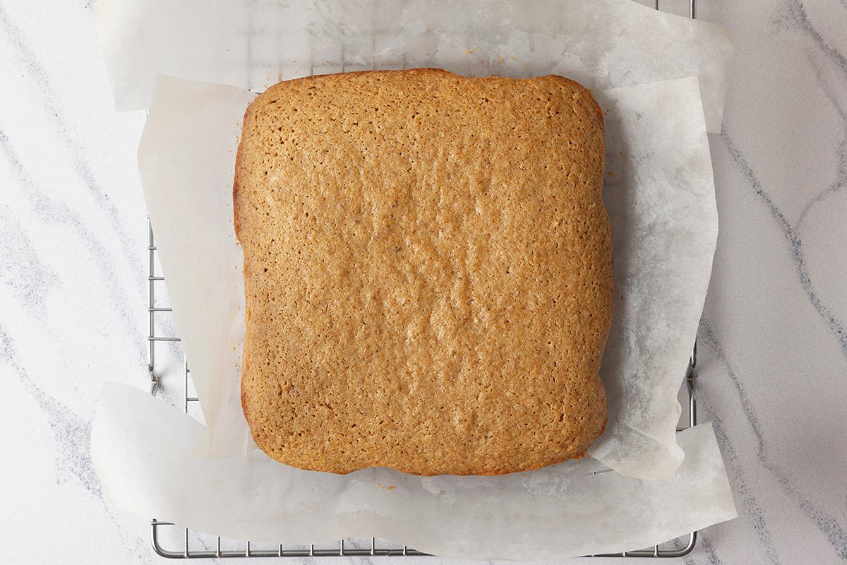 A square gingerbread sheet cake cooling on a wire rack, lined with parchment paper. The cake has a golden-brown color and sits on a light marble surface.