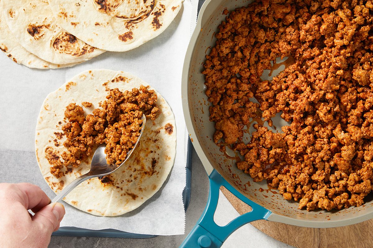 overhead shot of a scoop of seasoned ground meat is being placed onto a flat, round tortilla, which is positioned on a sheet of parchment paper; next to this, there is a pan filled with more of the cooked ground meat