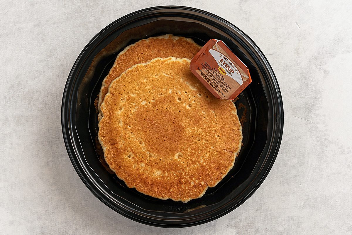 A black plate with two golden-brown pancakes. On top, there is a small unopened container of butter. The background is a light, textured surface.