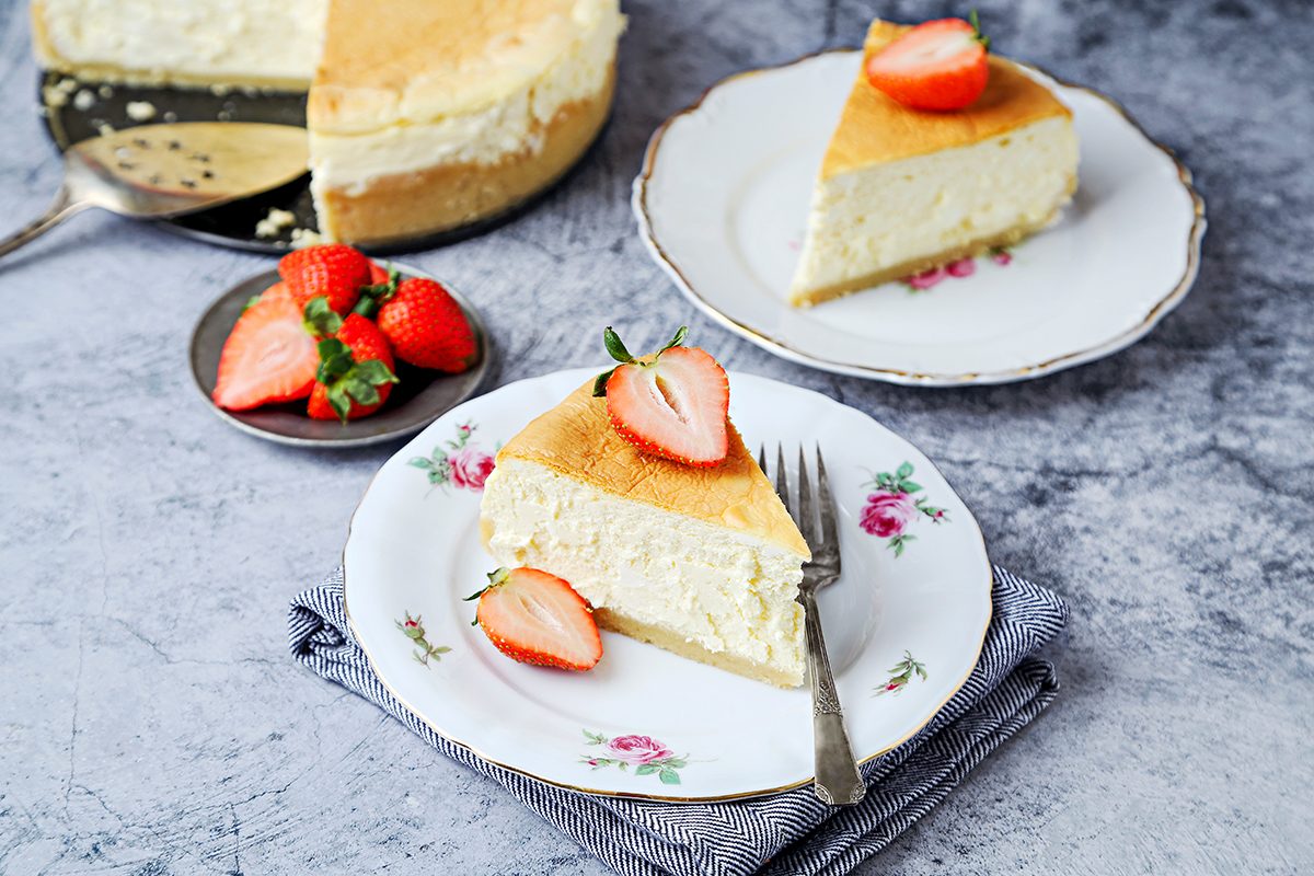 A slice of cheesecake topped with a halved strawberry on a floral plate with a fork rests on a fabric napkin. In the background, a full cheesecake and more strawberries are visible.