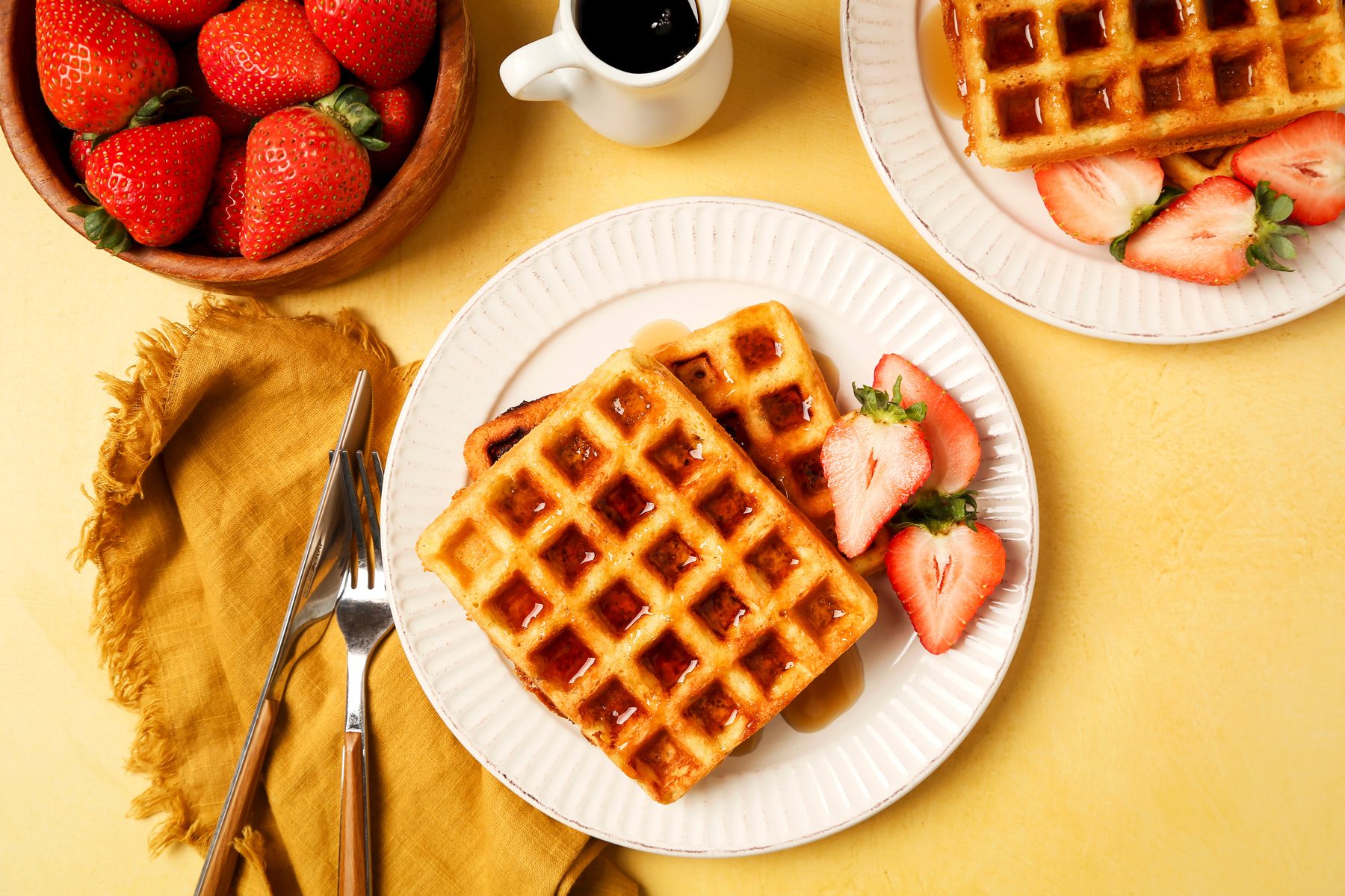Keto Waffles shot on a yellow surface. Waffles served on a white plate with syrup and strawberries.