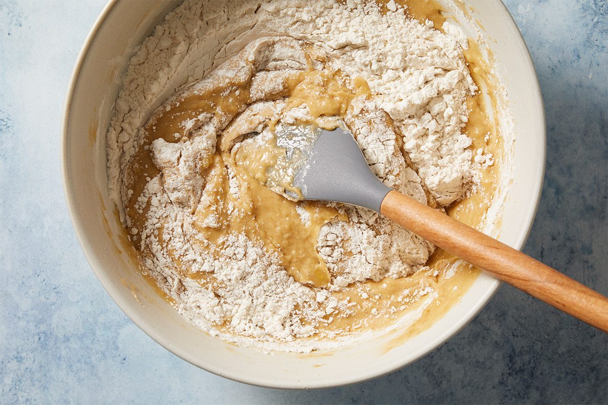 overhead shot of a mixing bowl filled with a mixture of flour and a yellowish batter; the batter is partially blended with the flour, a gray spatula with a wooden handle sits in the mixture, the bowl is placed against a light blue background