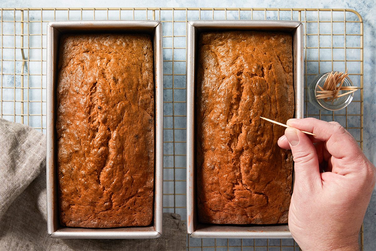 overhead shot of two freshly baked loaves placed side by side in metallic baking pans; in the foreground, a hand is holding a toothpick above the left loaf, a small glass container filled with additional toothpicks is visible nearby, along with a textured linen towel draped to one side; the background features a grid cooling rack on a light blue surface