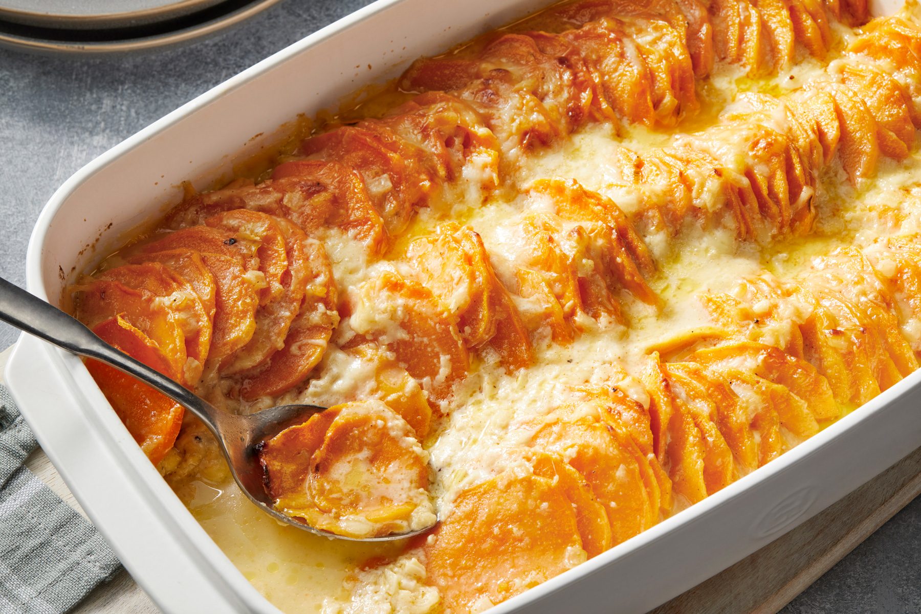 Close view shot of Savory Scalloped Sweet Potatoes; in a baking pan with serving spoon in it; over wooden board; with napkin and empty plates are nearby; all set on a grey surface;