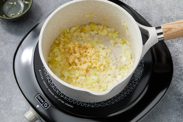 Overhead shot of a small saucepan; melt butter over medium heat; add onion; cook until tender 4-5 minutes; add garlic; cook one additional minute longer; over induction; all set on a grey surface;