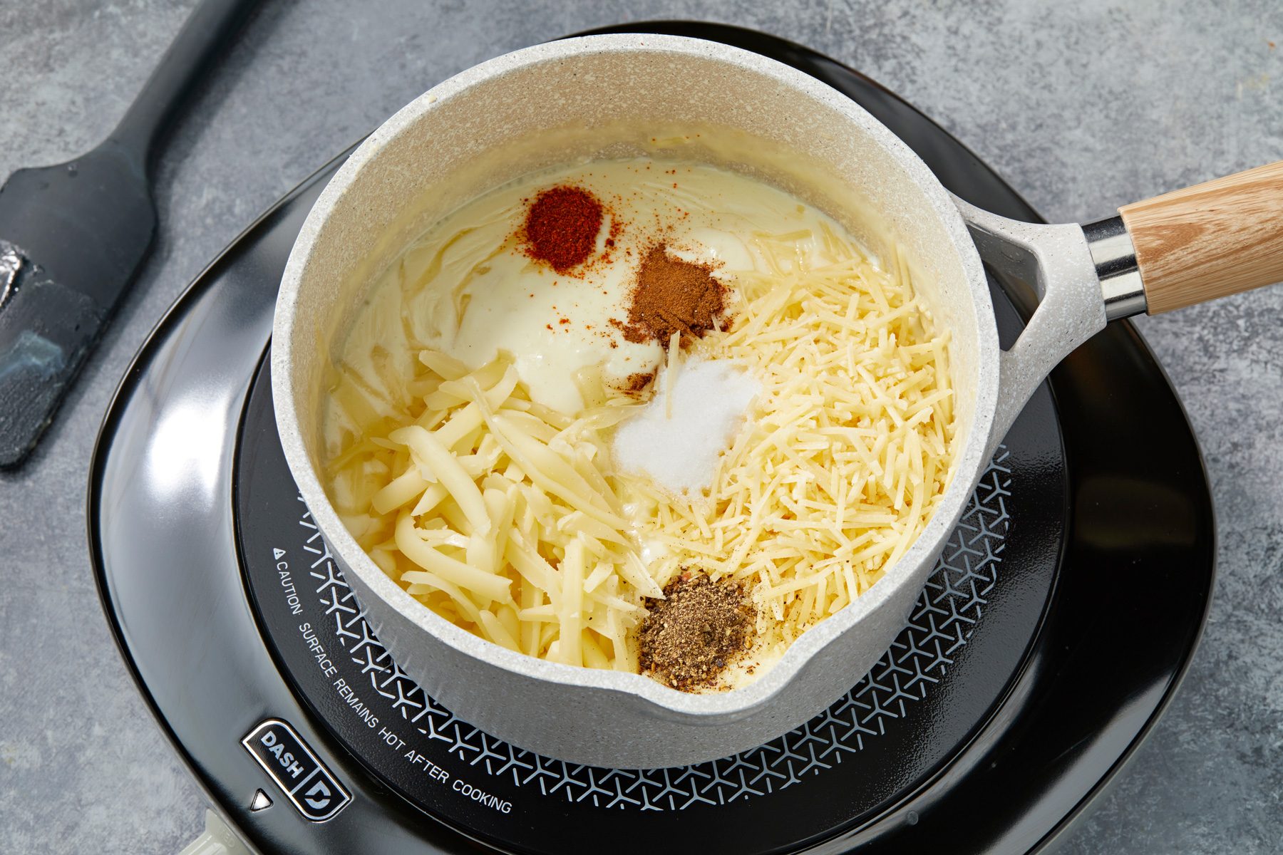 Overhead shot of the same saucepan; add salt; pepper; cayenne; cinnamon; Parmesan and 1 cup Gruyere; stir well; induction; spatula on a grey surface;