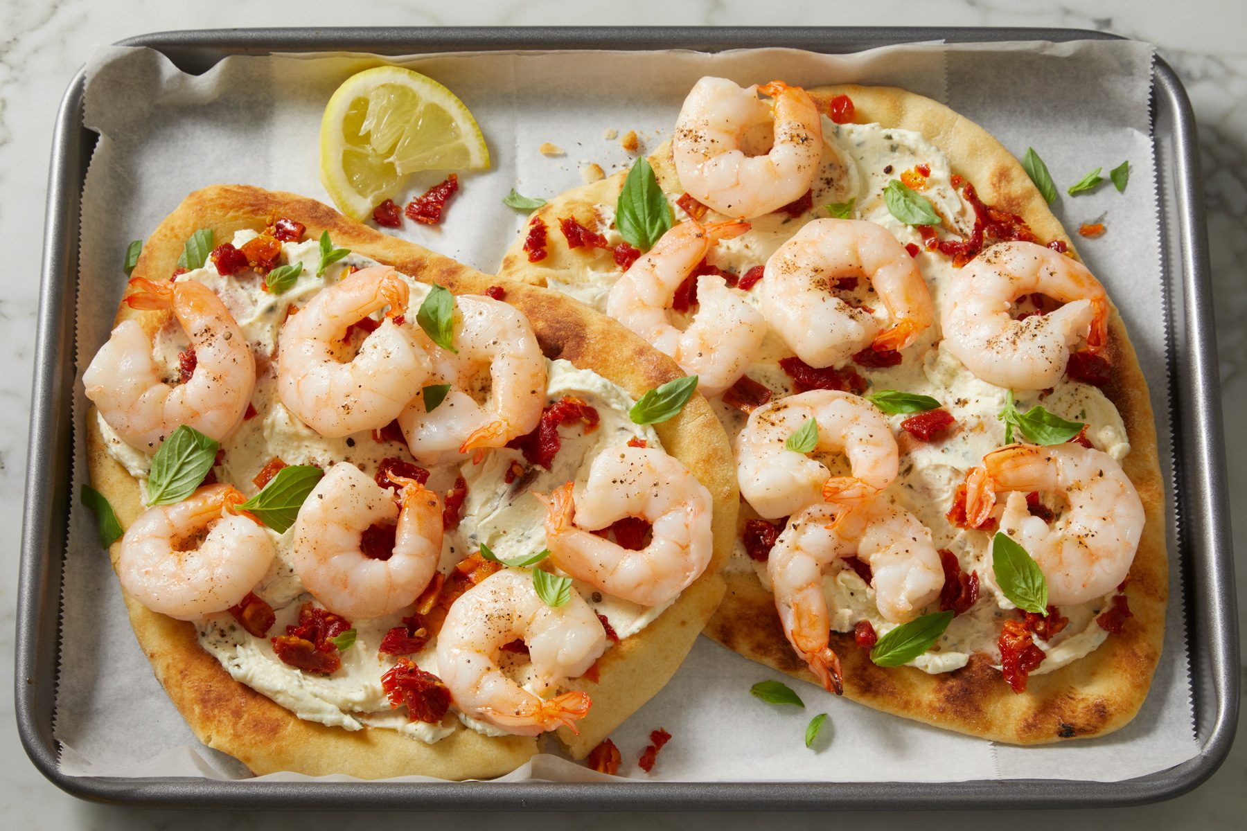 3/4th shot of Speedy Shrimp Flatbreads on a baking tray lined with parchment paper; a slice of lemon sits on the edge of the tray