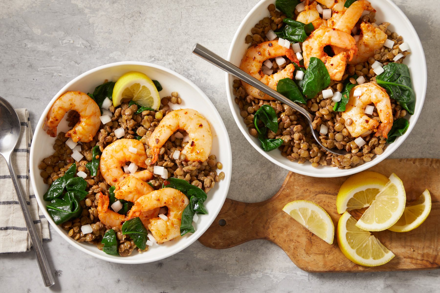 overhead shot of East Coast Shrimp and Lentil Bowls; scattered around the bowls are small pieces of lemon slices over wooden cutting board, the bowls are placed on a textured gray surface, a silver spoon rests beside one of the bowls