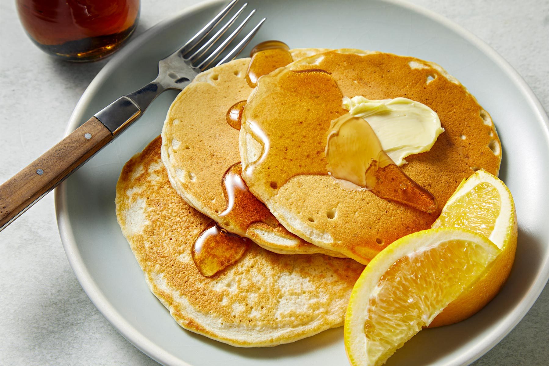 3/4th shot of sourdough pancakes served with a steel fork on a ceramic plate with butter, lemons and honey.