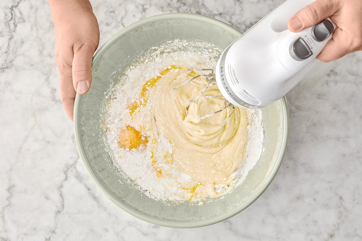 Hands holding a bowl while mixing cake batter with an electric hand mixer. The batter includes flour, eggs, and other ingredients on a marble countertop.